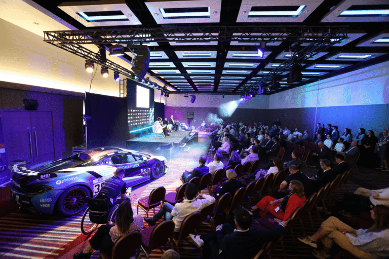 Audience facing a blue-lit stage and large presentation screens at the BlackBook Motorsport Forum.